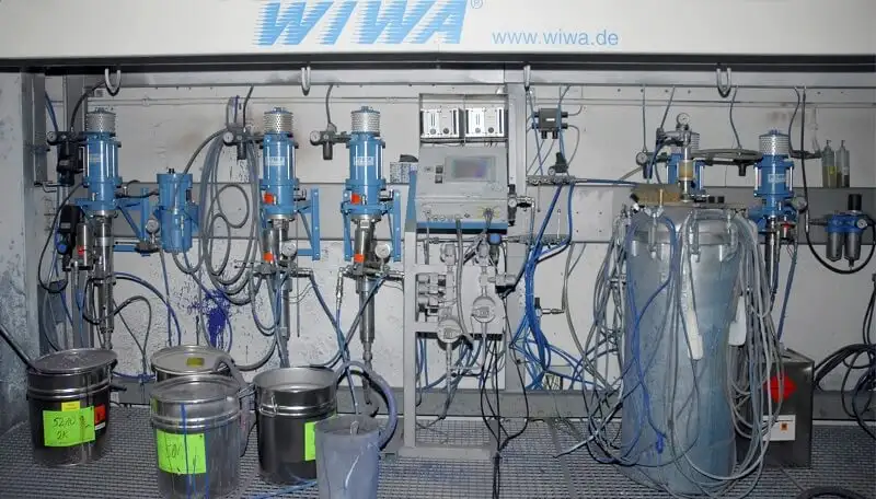 Окрасочные насосы WIWA с дополнительным оборудованием в производственном помещении. На стене установлены несколько насосов, система управления, датчики и многочисленные шланги. Спереди стоят ведра и контейнеры с краской, отмеченные цветными наклейками.