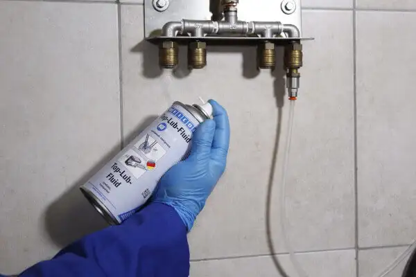 Человек в синей перчатке держит баллончик смазки WEICON Top-Lub Fluid перед соединением сжатого воздуха на плитке.