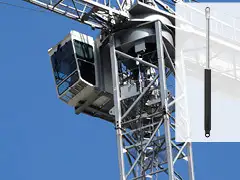 Газовий силовий привід, встановлений на дверях кабіни баштового крана. На фоні видно металеву конструкцію крана та кабіну оператора, що розташована на великій висоті. Праворуч показано окремий силовий привід із чорним корпусом і металевим штоком для плавного відкривання та утримання дверей.        Запитати в ChatGPT