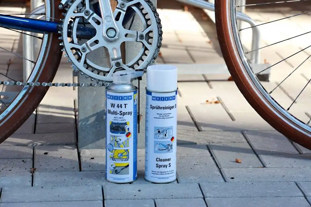 Два балони (W 44 T Multi-Spray та Cleaner Spray S) для чищення та догляду за велосипедним ланцюгом та зірочками. Засоби стоять на бруківці на тлі чистого ланцюга та колеса велосипеда. Фото ілюструє консервацію та обслуговування трансмісії.