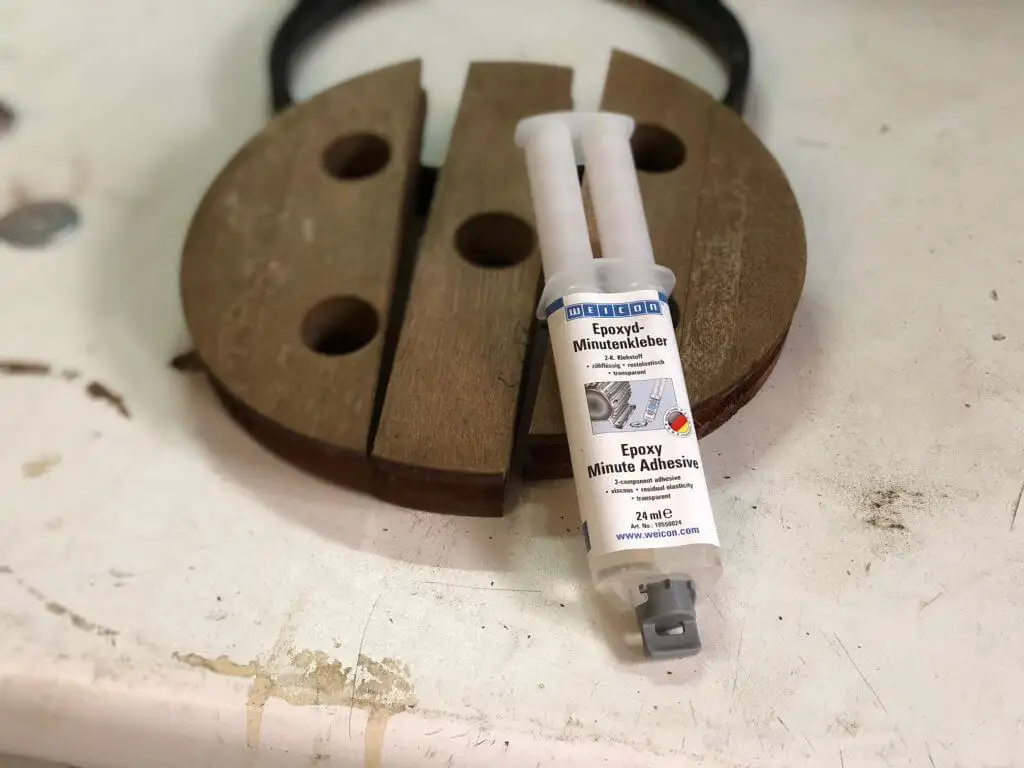 Двокомпонентний епоксидний клей Weicon "Epoxyd Minute Adhesive" у шприці, розташований поруч із розколотим дерев'яним елементом (диском) від садового лежака. Клей використовується для швидкого та міцного ремонту.