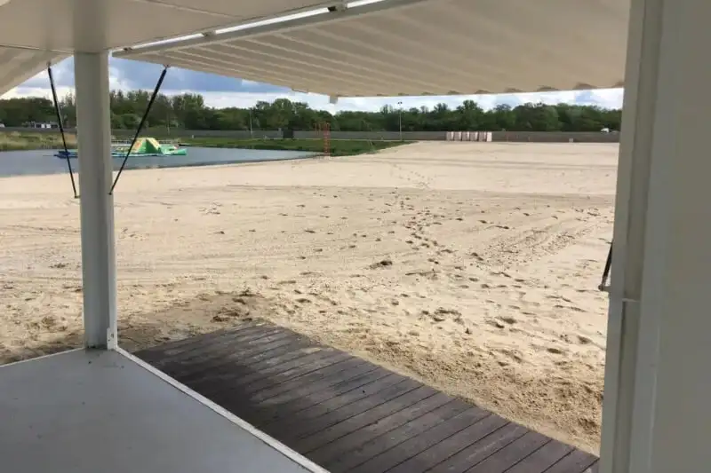 Панорамний вид на піщаний пляж з водоймою та зеленню, знятий зсередини білого контейнера або павільйону, з дерев'яним настилом на передньому плані.