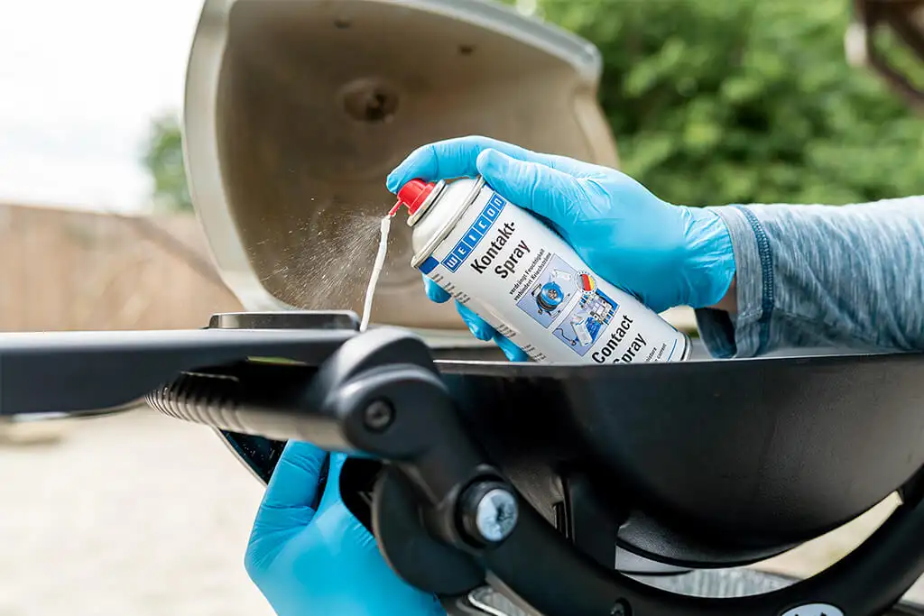 Руки в синіх рукавичках використовують спрей Weicon Kontakt-Spray для чищення електричних контактів та елементів запалювання газового гриля.