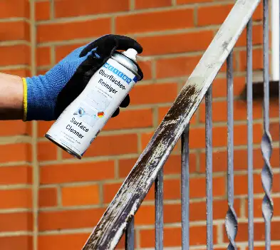 Рука в робочій рукавичці розпилює очисник поверхонь (Surface Cleaner) на забруднені металеві перила перед видаленням іржі.