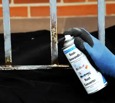 Рука в рукавичці наносить спрей-перетворювач іржі (Rust Converter) на іржаві металеві ґрати.