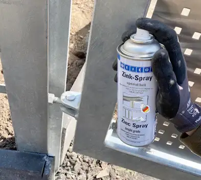 Рука в рукавичці наносить яскраво-цинковий спрей Weicon (Zinc Spray bright grade) для захисту нової металевої садової брами від корозії.