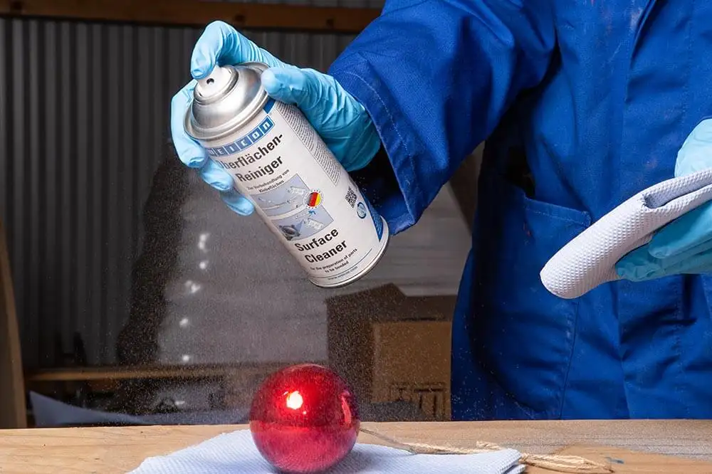 Людина в синіх рукавичках та робочому одязі розпилює WEICON Surface Cleaner (Очисник поверхонь) на червону глянцеву ялинкову кульку, підготовлюючи її поверхню до фарбування або оновлення