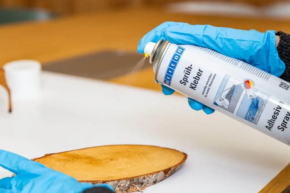Людина в синіх захисних рукавичках наносить клей-спрей WEICON Adhesive Spray на поверхню овального зрізу березового дерева. Процес підготовки основи для різдвяного декору своїми руками